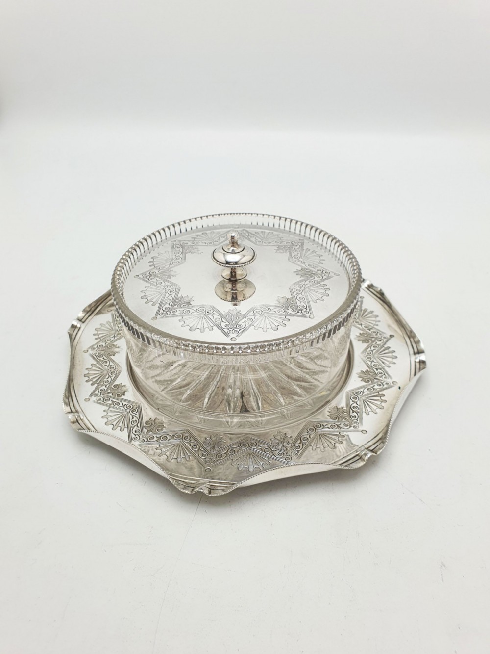 A Victorian silver-plated circular butter dish featuring bright-cut engraving and a lid with a central finial, housing a clear cut-glass liner.