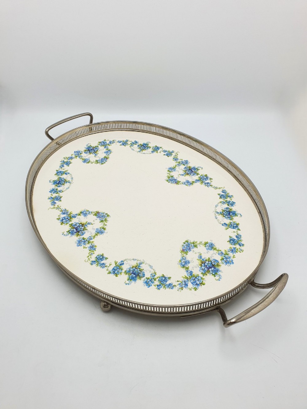 An antique oval serving tray featuring a pierced metal gallery rim, side handles, and an off-white ceramic base decorated with a blue floral garland.