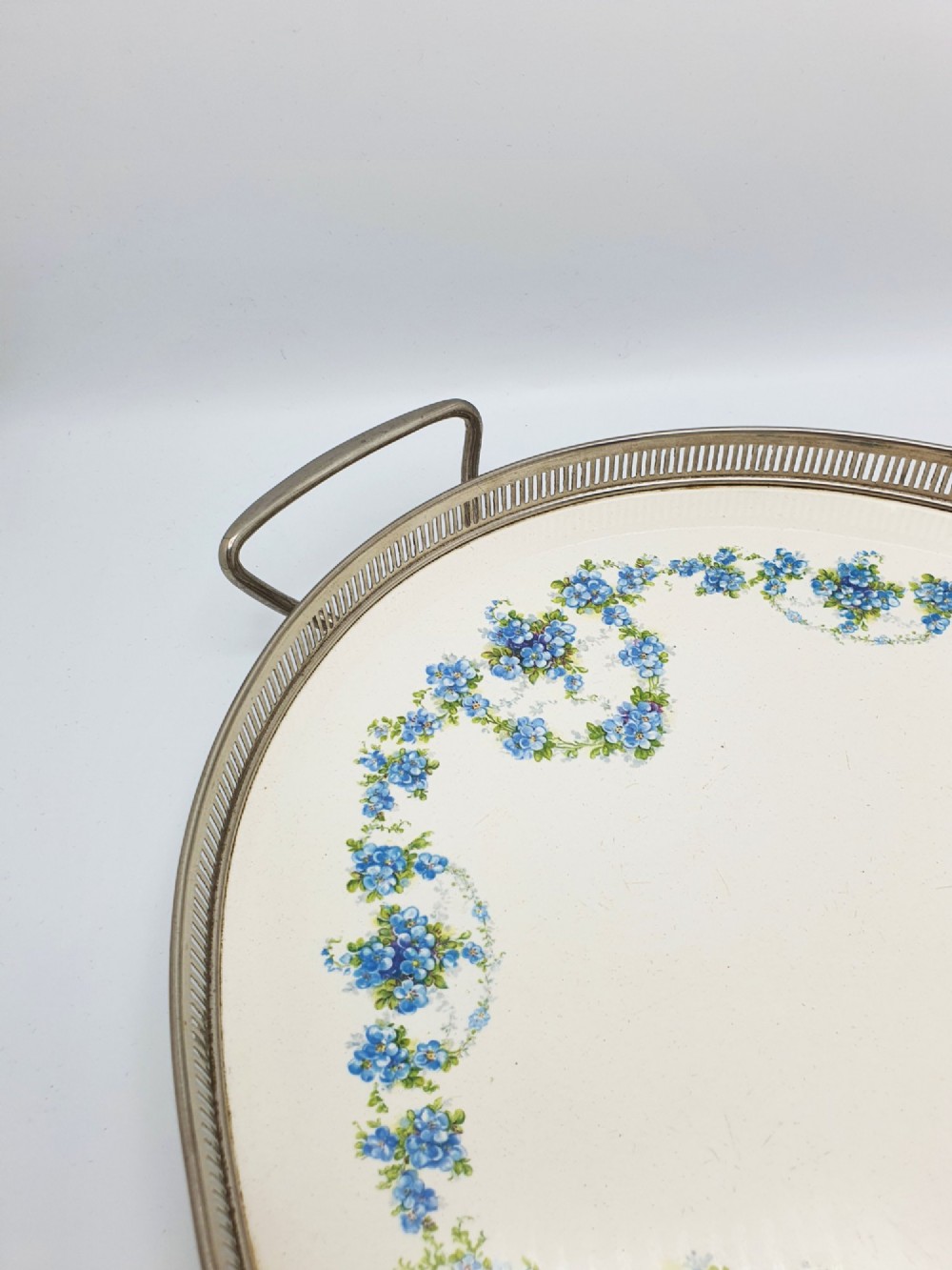 An antique oval serving tray featuring a pierced metal gallery rim, side handles, and an off-white ceramic base decorated with a blue floral garland. View II