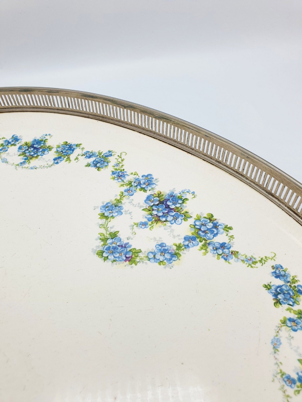 An antique oval serving tray featuring a pierced metal gallery rim, side handles, and an off-white ceramic base decorated with a blue floral garland. View III