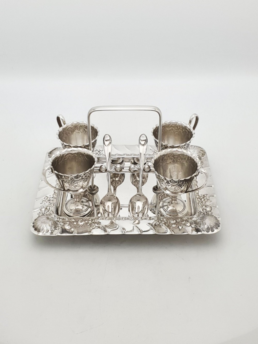 An Edwardian silver-plated egg cruet set with four embossed egg cups, four spoons, and an ornate tray with a central carry handle. View II