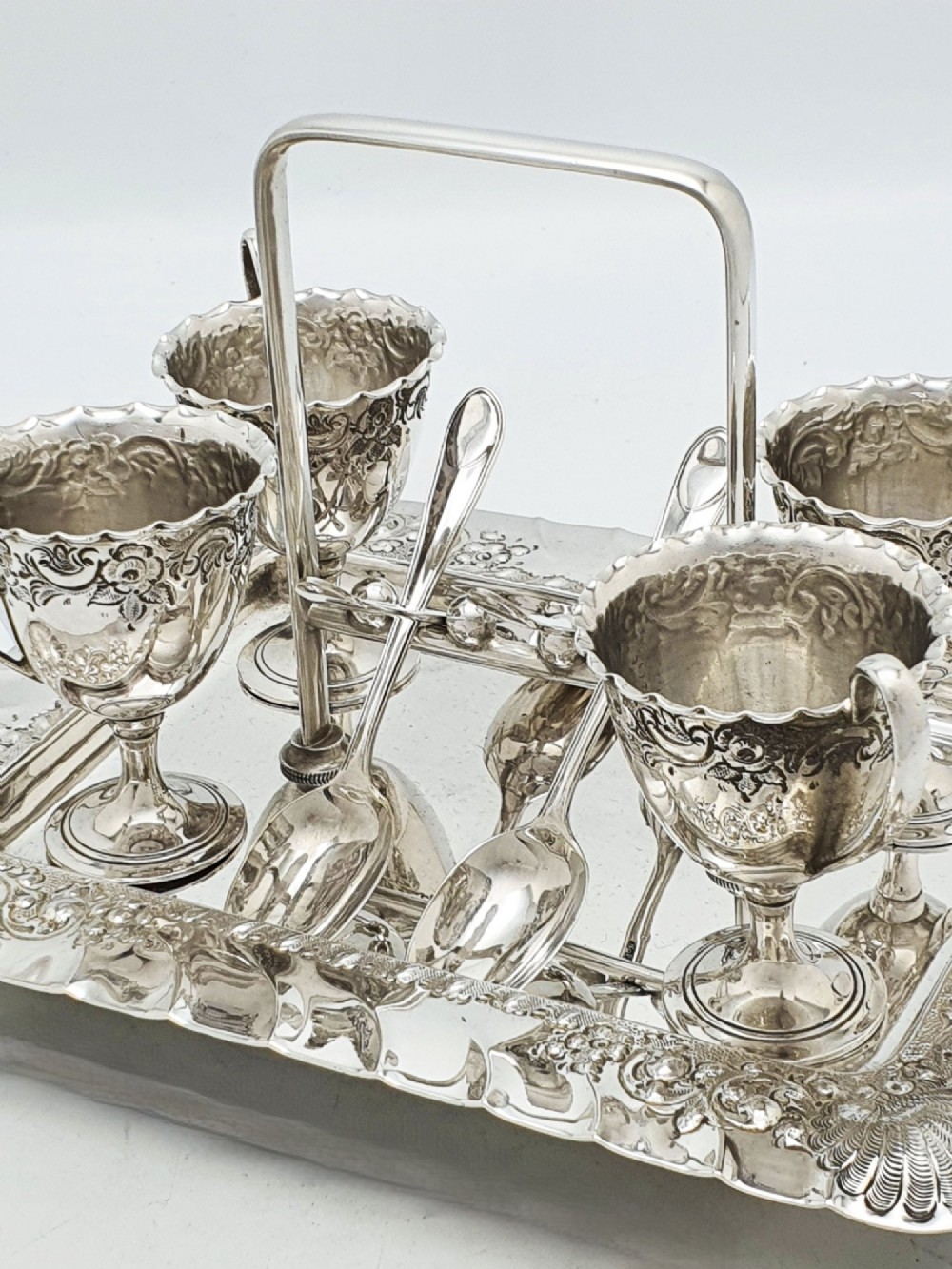An Edwardian silver-plated egg cruet set with four embossed egg cups, four spoons, and an ornate tray with a central carry handle. View V