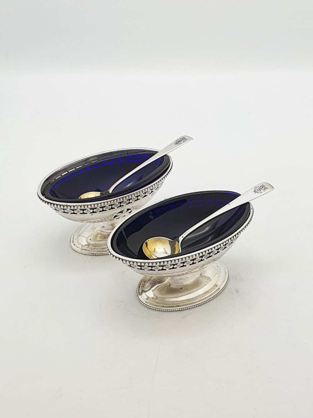 A pair of Edwardian sterling silver oval pedestal salt cellars with pierced filigree rims, dark cobalt blue glass liners, and matching silver salt spoons.
