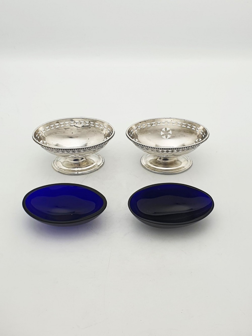 A pair of Edwardian sterling silver oval pedestal salt cellars with pierced filigree rims, dark cobalt blue glass liners, and matching silver salt spoons. View IV