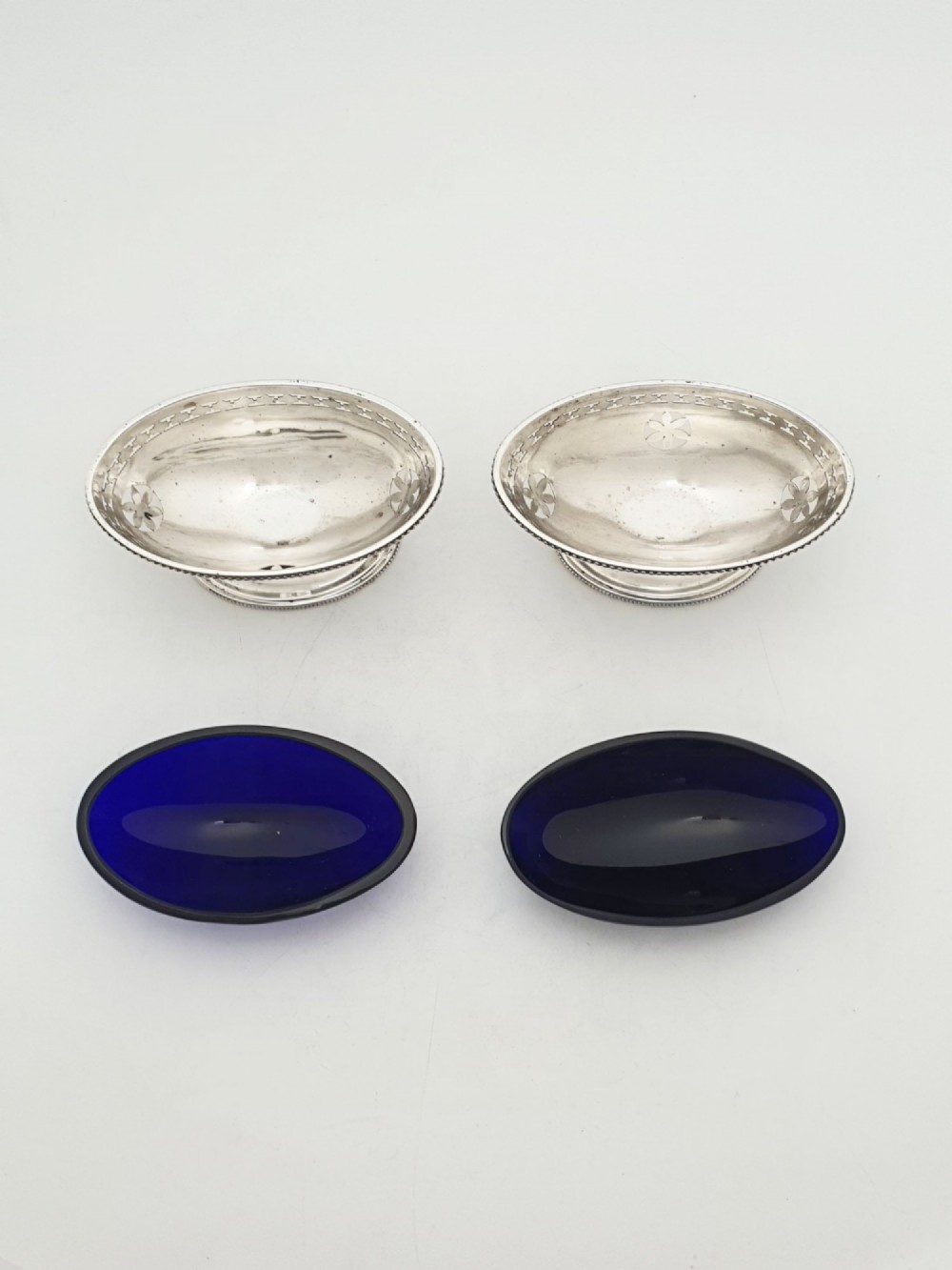 A pair of Edwardian sterling silver oval pedestal salt cellars with pierced filigree rims, dark cobalt blue glass liners, and matching silver salt spoons. View V