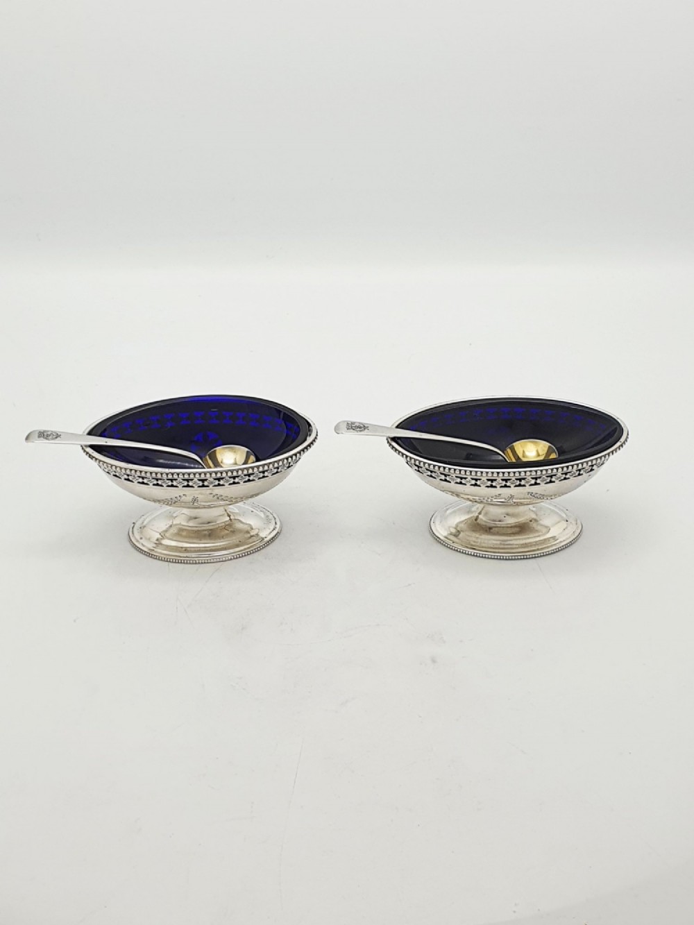 A pair of Edwardian sterling silver oval pedestal salt cellars with pierced filigree rims, dark cobalt blue glass liners, and matching silver salt spoons. View IX