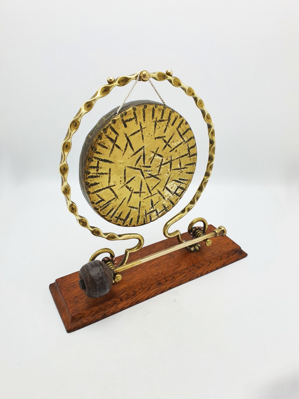 An antique brass circular gong with patterned etching suspended within a wavy brass frame on a polished rectangular oak base with a matching striker.