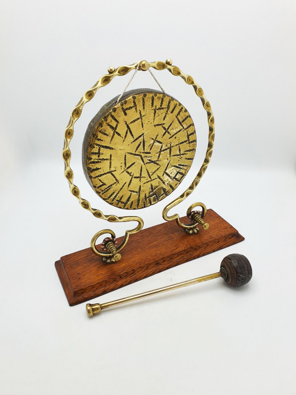 An antique brass circular gong with patterned etching suspended within a wavy brass frame on a polished rectangular oak base with a matching striker. View II