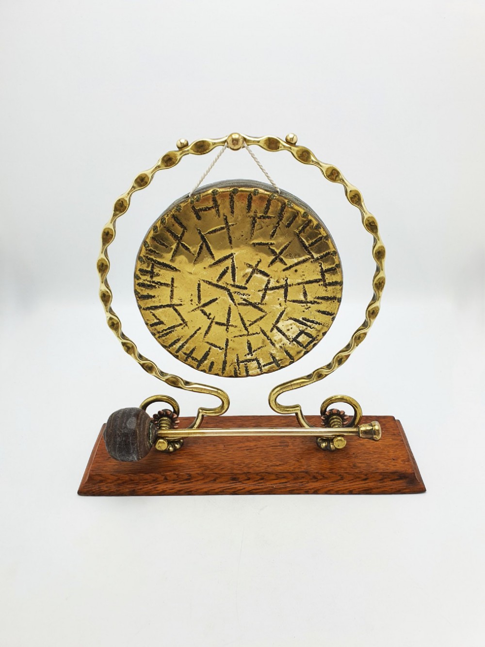 An antique brass circular gong with patterned etching suspended within a wavy brass frame on a polished rectangular oak base with a matching striker. View III