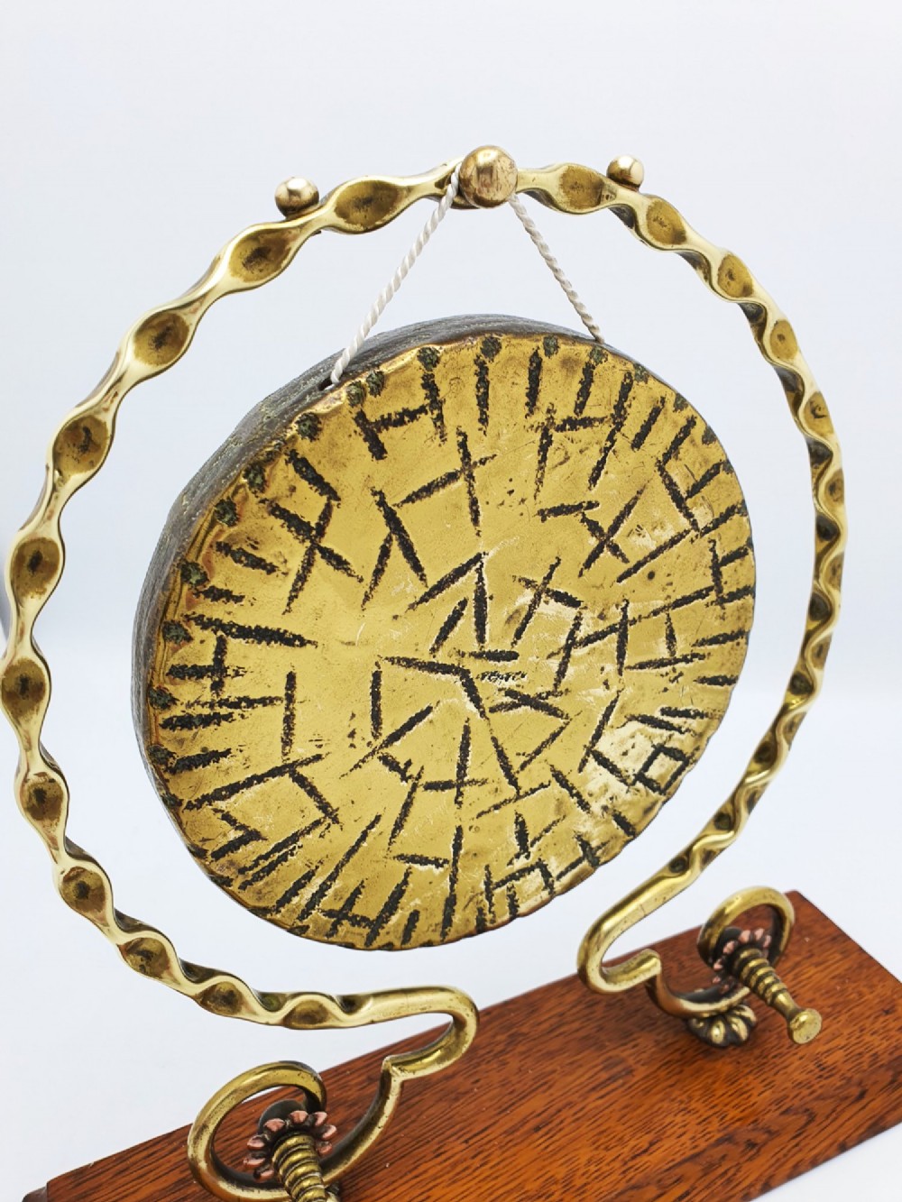 An antique brass circular gong with patterned etching suspended within a wavy brass frame on a polished rectangular oak base with a matching striker. View V