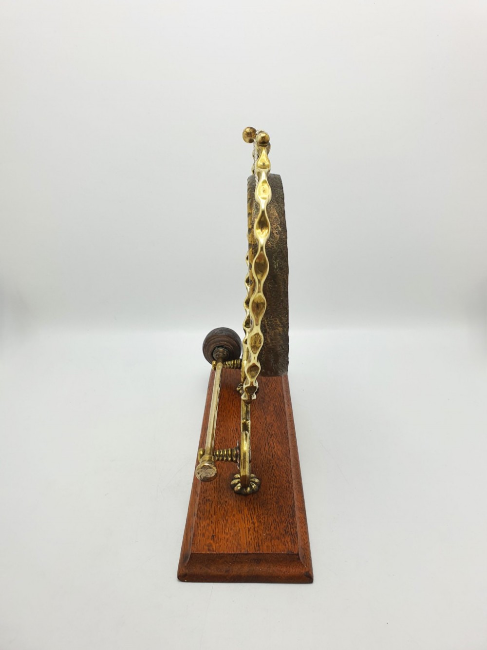 An antique brass circular gong with patterned etching suspended within a wavy brass frame on a polished rectangular oak base with a matching striker. View VI