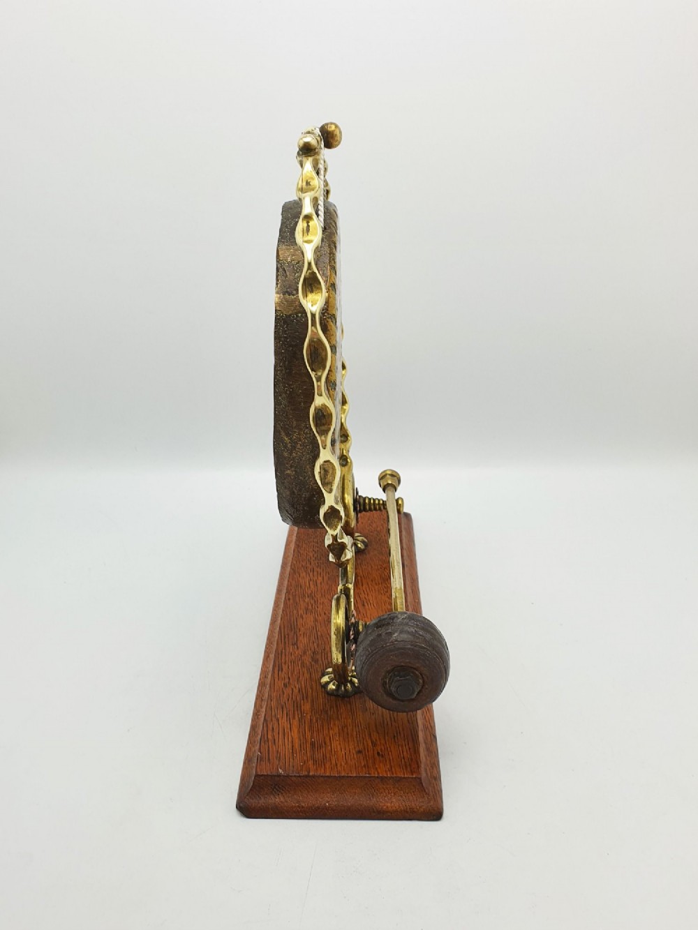 An antique brass circular gong with patterned etching suspended within a wavy brass frame on a polished rectangular oak base with a matching striker. View VIII