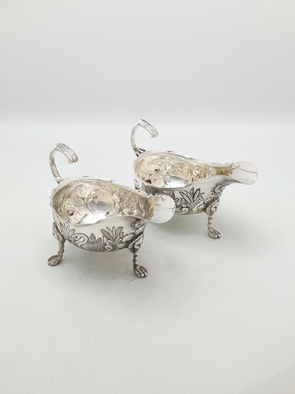A pair of engraved silver-plated sauce boats with curved handles and decorated cabriole legs resting on a neutral background.