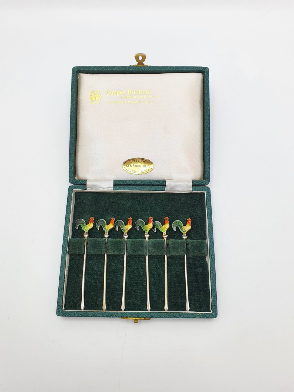 A set of six silver cocktail sticks featuring colourful enamelled cockerel finials arranged inside a dark green presentation box.