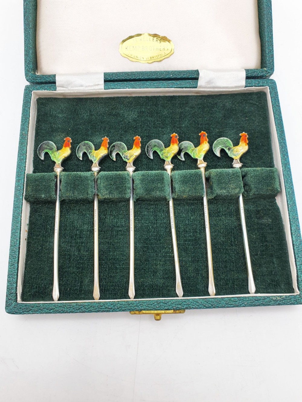 A set of six silver cocktail sticks featuring colourful enamelled cockerel finials arranged inside a dark green presentation box. View II