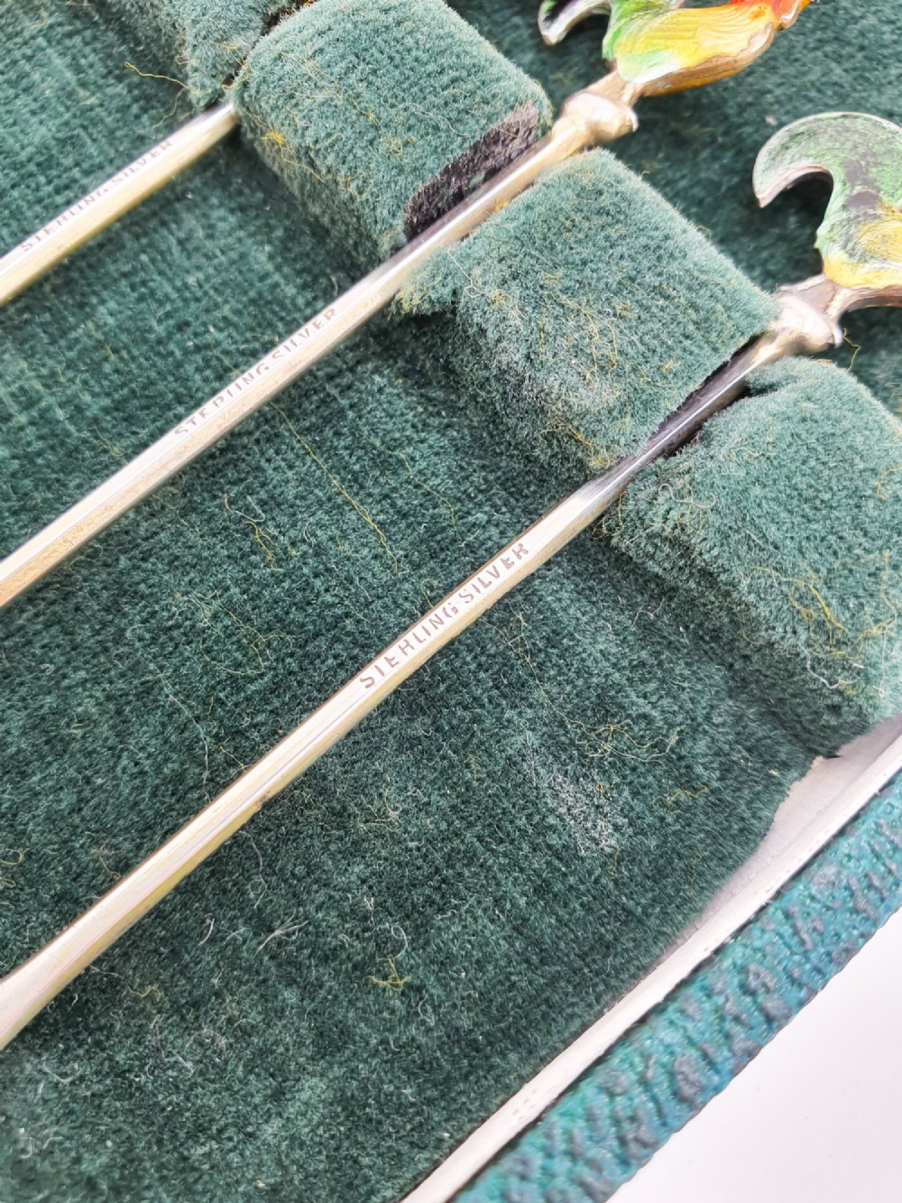 A set of six silver cocktail sticks featuring colourful enamelled cockerel finials arranged inside a dark green presentation box. View III