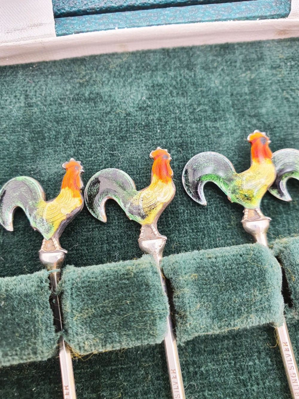 A set of six silver cocktail sticks featuring colourful enamelled cockerel finials arranged inside a dark green presentation box. View IV