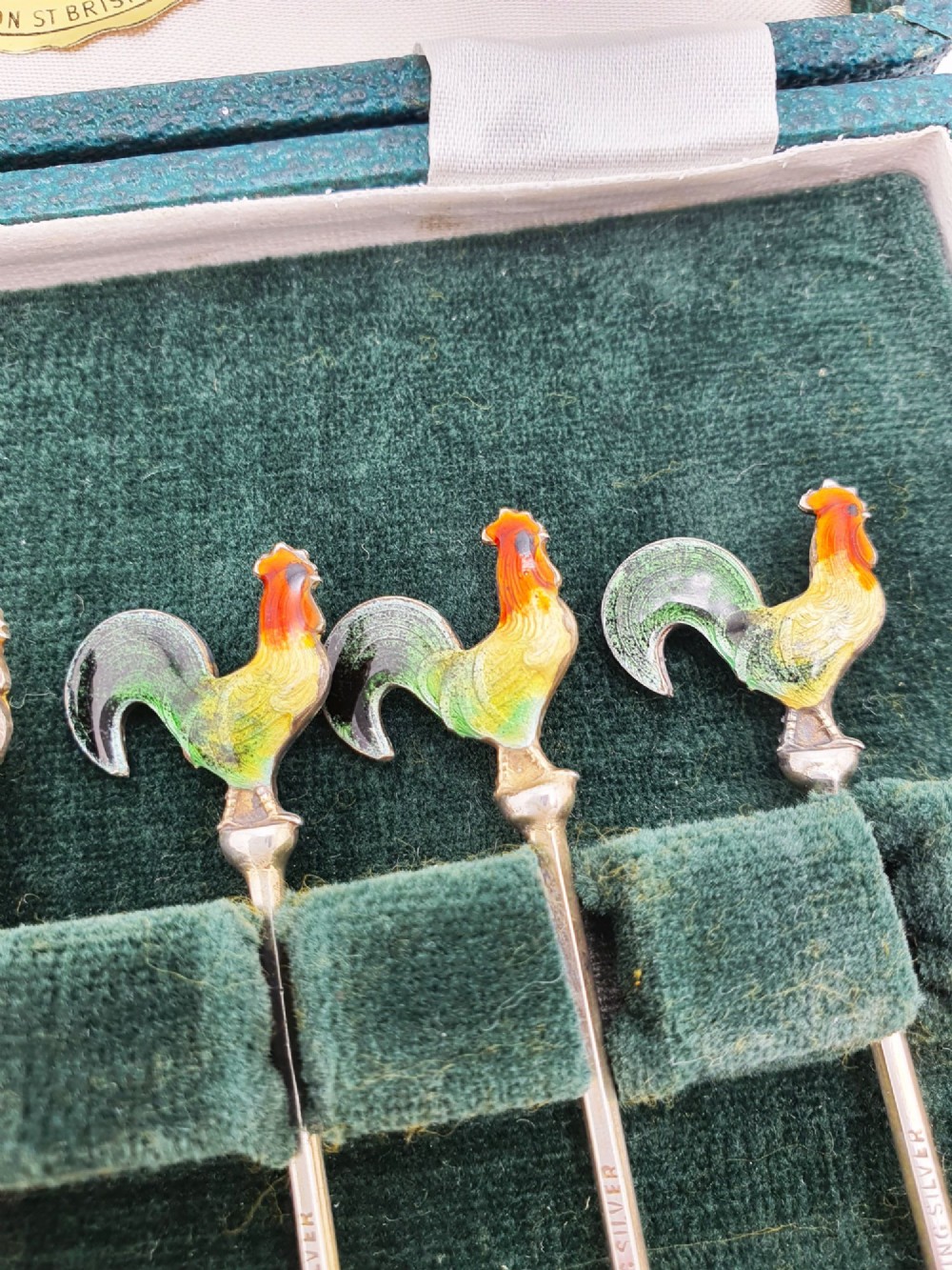 A set of six silver cocktail sticks featuring colourful enamelled cockerel finials arranged inside a dark green presentation box. View V