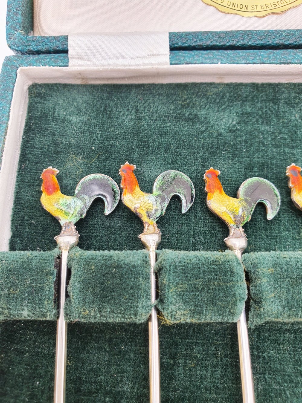 A set of six silver cocktail sticks featuring colourful enamelled cockerel finials arranged inside a dark green presentation box. View VI