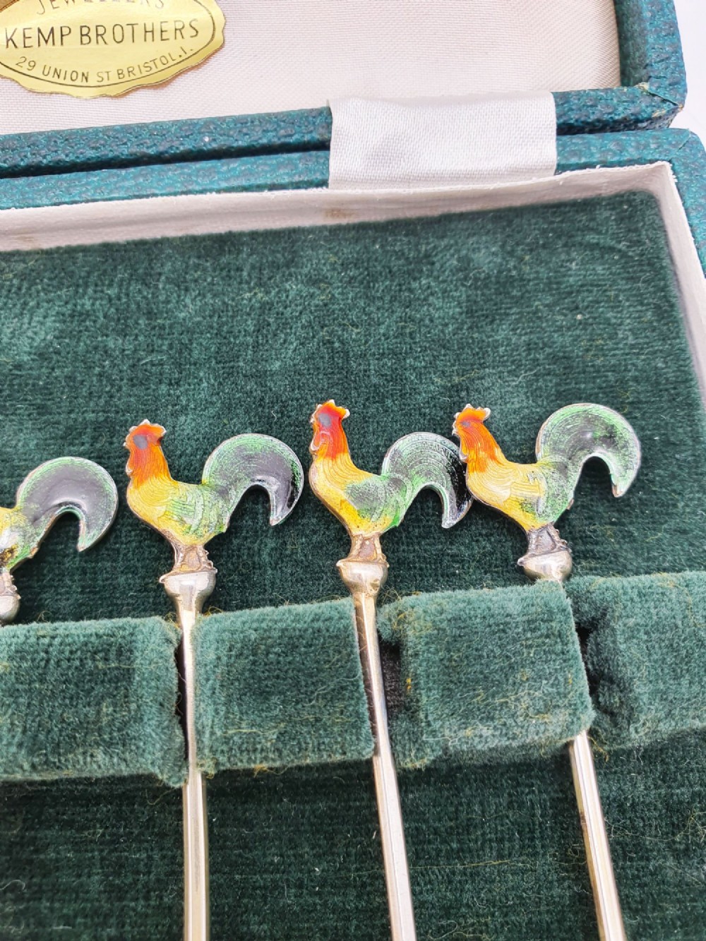 A set of six silver cocktail sticks featuring colourful enamelled cockerel finials arranged inside a dark green presentation box. View VII