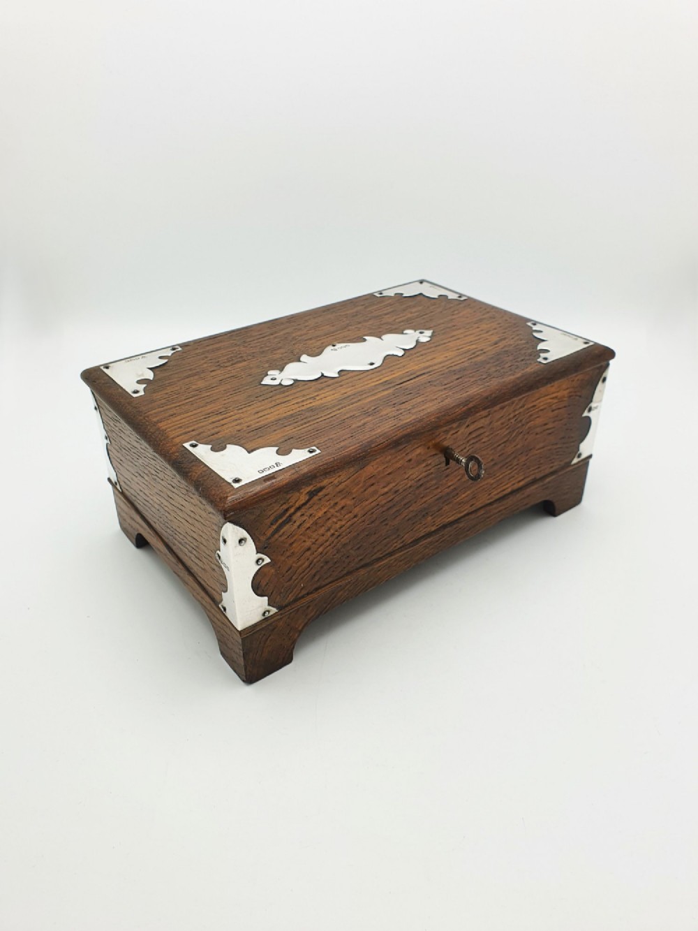 An Edwardian oak cigar box featuring decorative sterling silver corner mounts, a central shaped silver plaque, and a front keyhole with key.