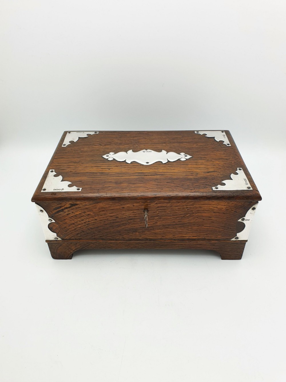 An Edwardian oak cigar box featuring decorative sterling silver corner mounts, a central shaped silver plaque, and a front keyhole with key. View III