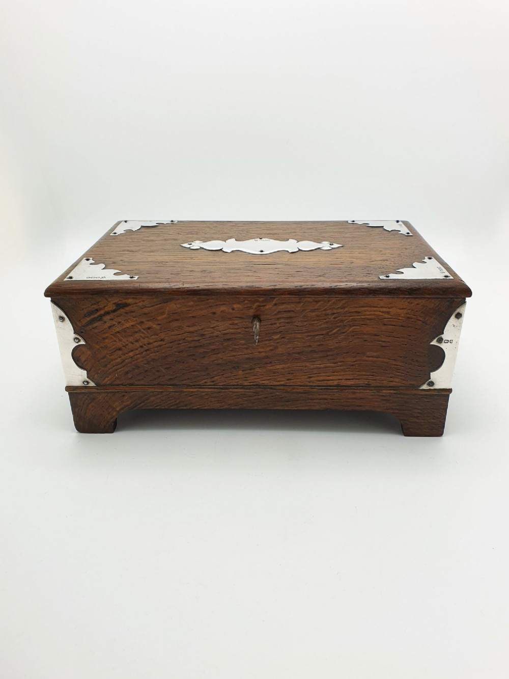 An Edwardian oak cigar box featuring decorative sterling silver corner mounts, a central shaped silver plaque, and a front keyhole with key. View VIII