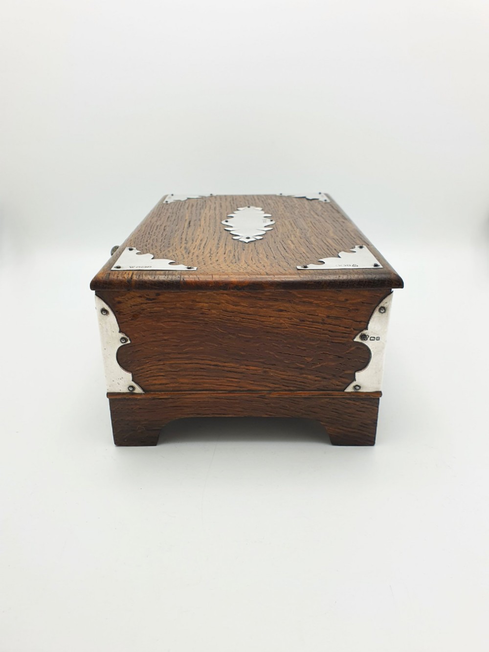 An Edwardian oak cigar box featuring decorative sterling silver corner mounts, a central shaped silver plaque, and a front keyhole with key. View IX