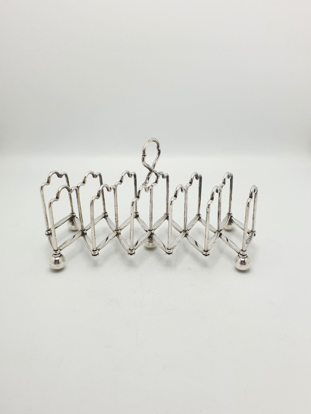 An antique silver-plated extending toast rack featuring an articulated concertina base, arched dividers, ball feet, and a central loop handle. View V