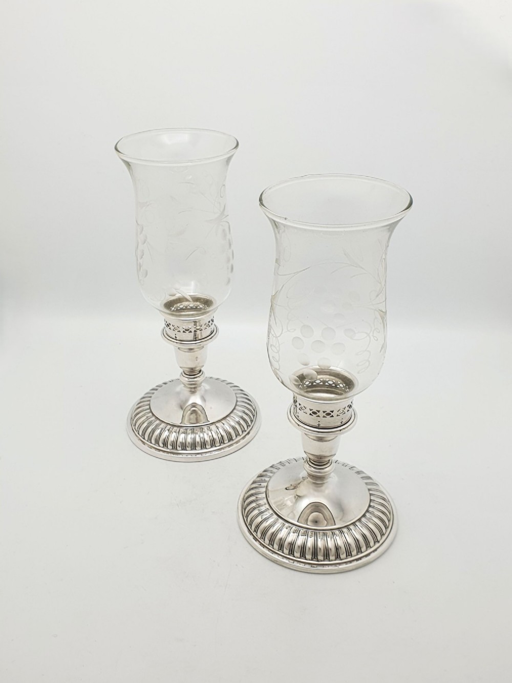 A pair of silver-plated candle holders featuring gadrooned bases and pierced sockets topped with etched glass hurricane shades.
