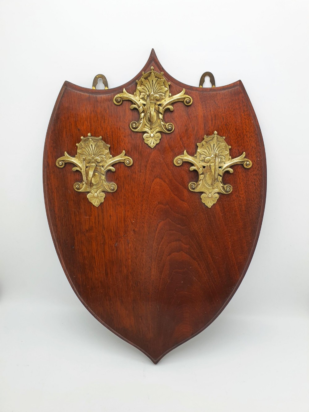 A shield-shaped mahogany wall plaque equipped with three decorative cast brass coat hooks arranged symmetrically.