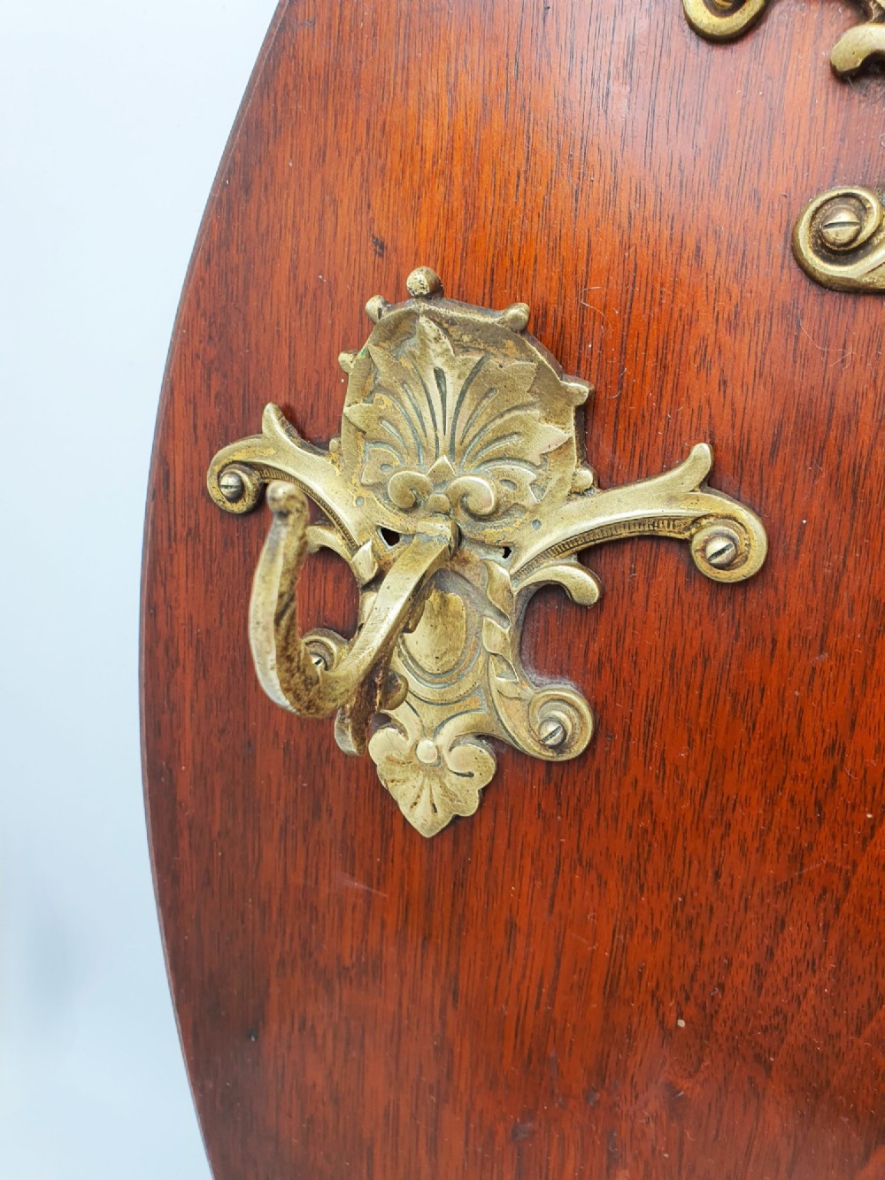 A shield-shaped mahogany wall plaque equipped with three decorative cast brass coat hooks arranged symmetrically. View IV
