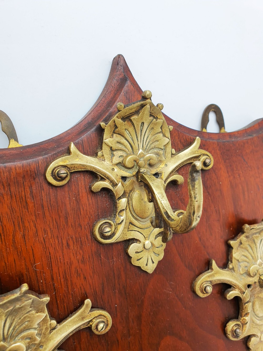 A shield-shaped mahogany wall plaque equipped with three decorative cast brass coat hooks arranged symmetrically. View V