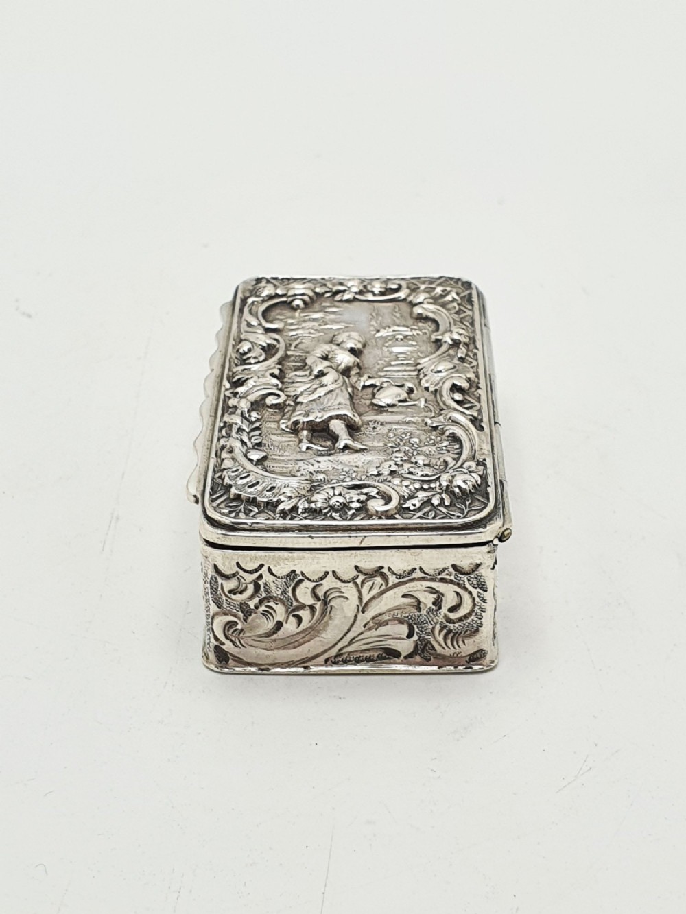 A rectangular Victorian sterling silver snuff box with intricate repousse relief work depicting a pastoral scene surrounded by floral scrollwork. View V