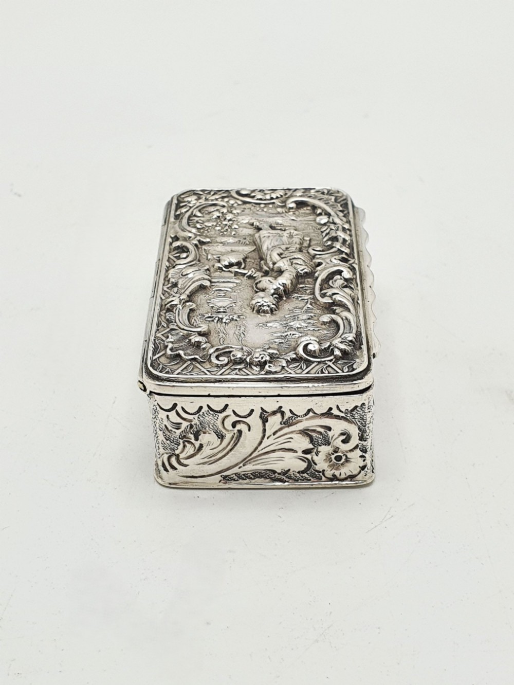 A rectangular Victorian sterling silver snuff box with intricate repousse relief work depicting a pastoral scene surrounded by floral scrollwork. View VII
