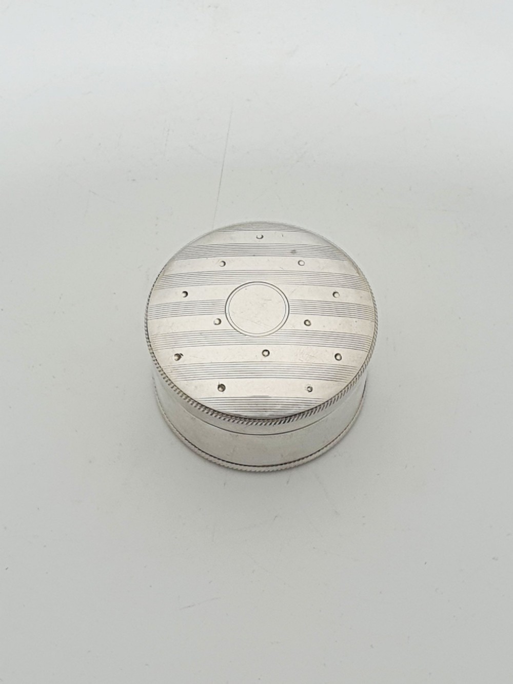 A circular sterling silver pill box featuring engine-turned linear engraving, small dots, and a central plain round cartouche.