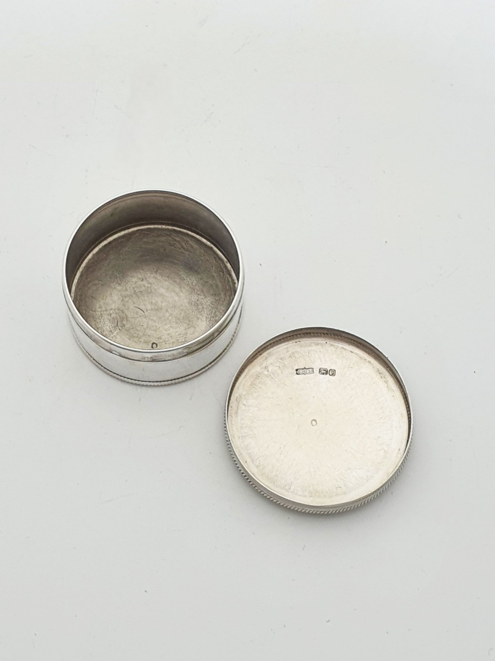 A circular sterling silver pill box featuring engine-turned linear engraving, small dots, and a central plain round cartouche. View II
