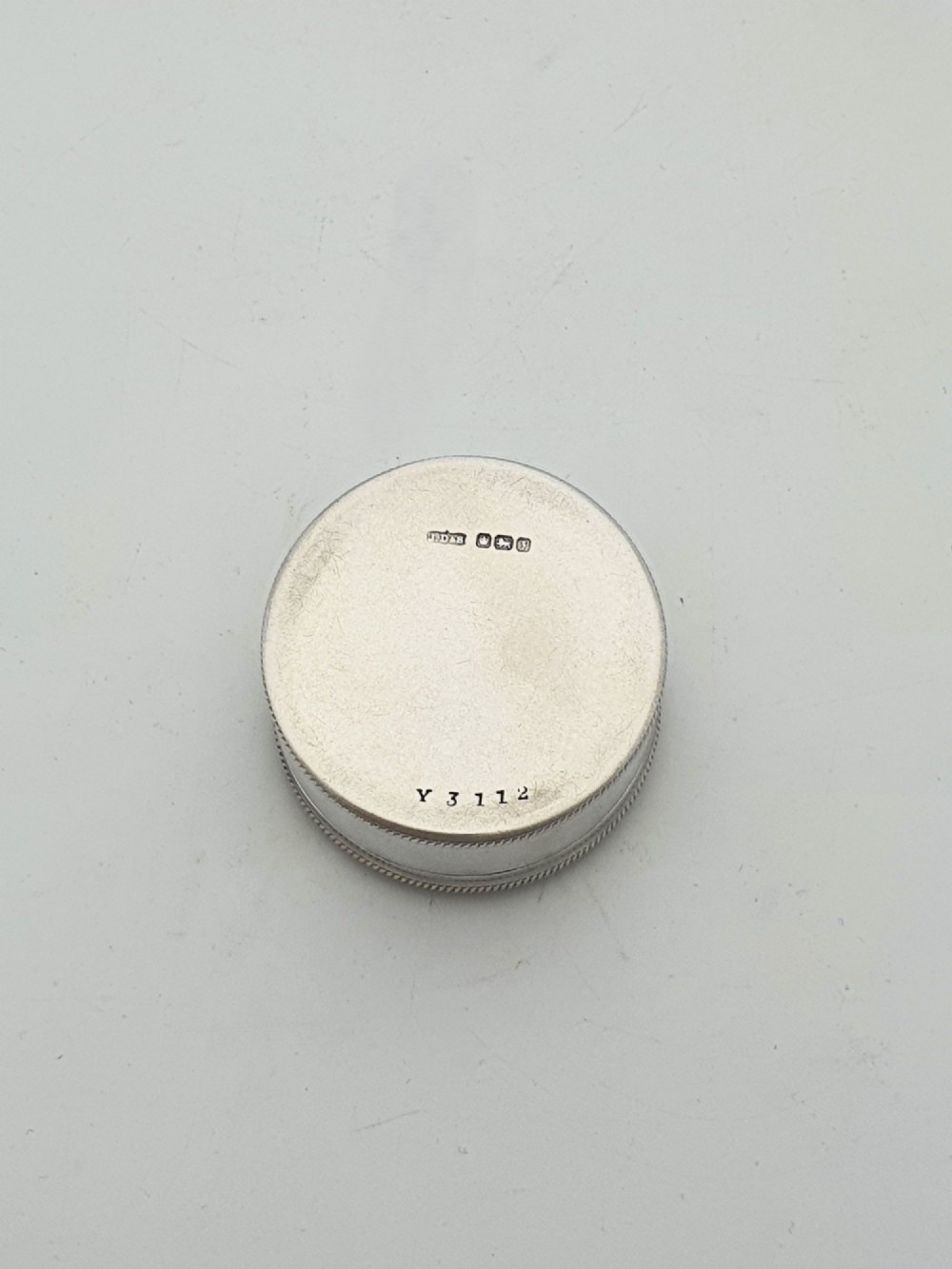 A circular sterling silver pill box featuring engine-turned linear engraving, small dots, and a central plain round cartouche. View III