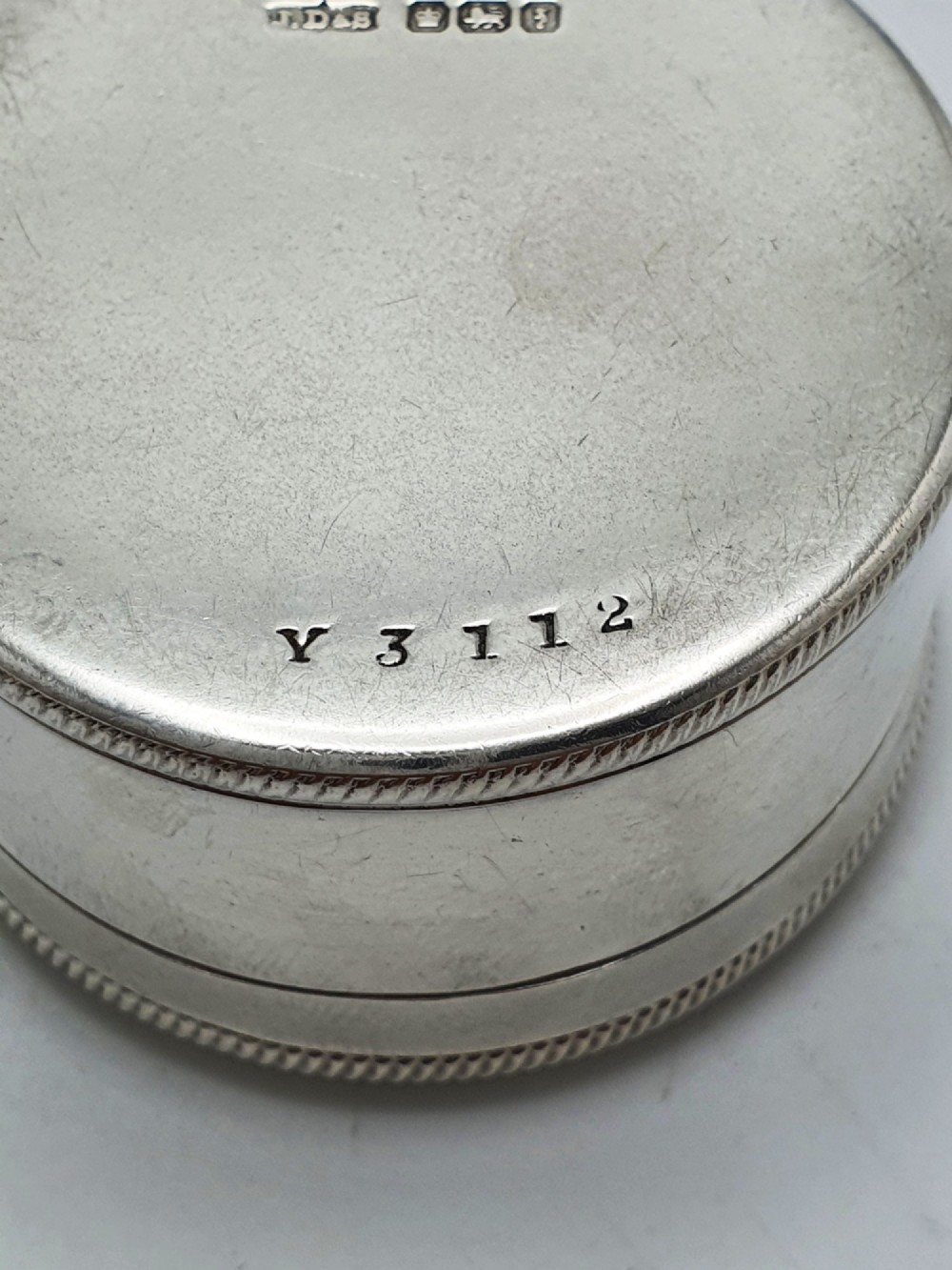 A circular sterling silver pill box featuring engine-turned linear engraving, small dots, and a central plain round cartouche. View V