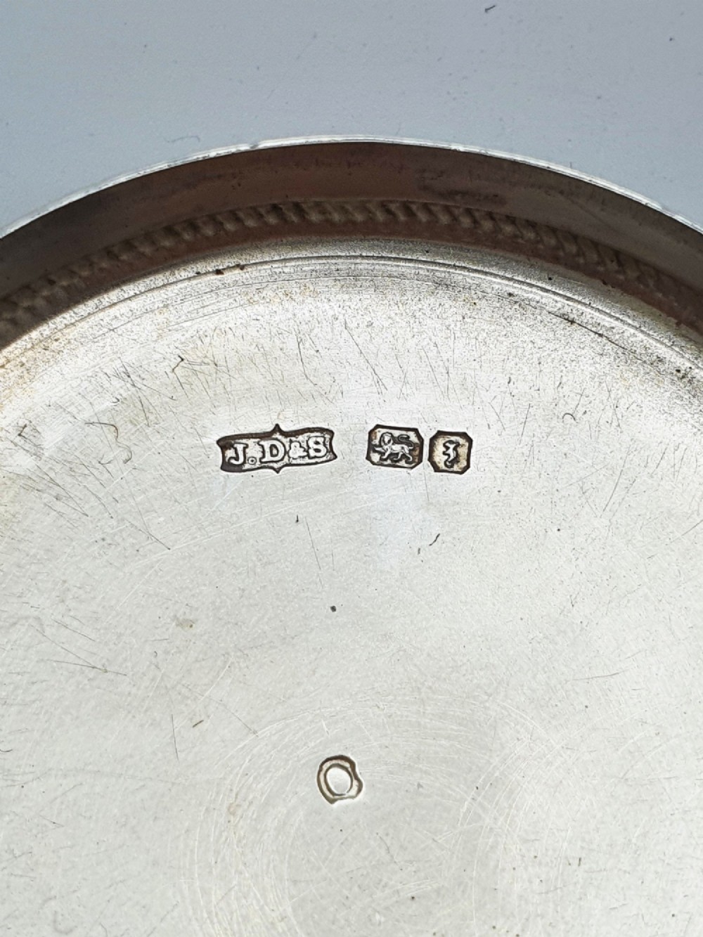 A circular sterling silver pill box featuring engine-turned linear engraving, small dots, and a central plain round cartouche. View VI