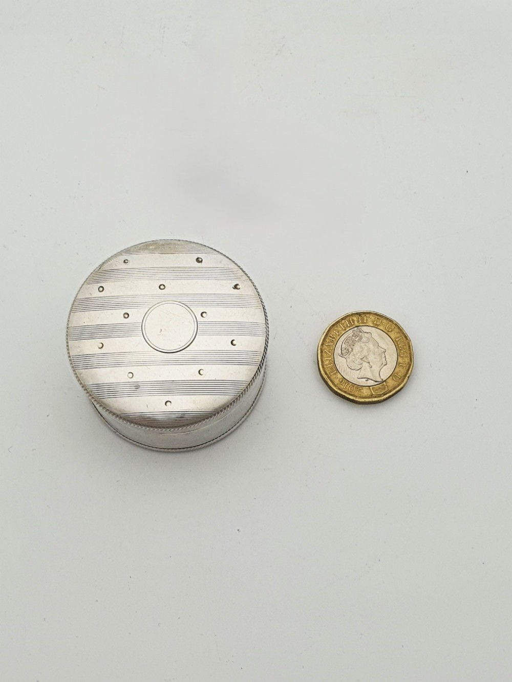 A circular sterling silver pill box featuring engine-turned linear engraving, small dots, and a central plain round cartouche. View VII