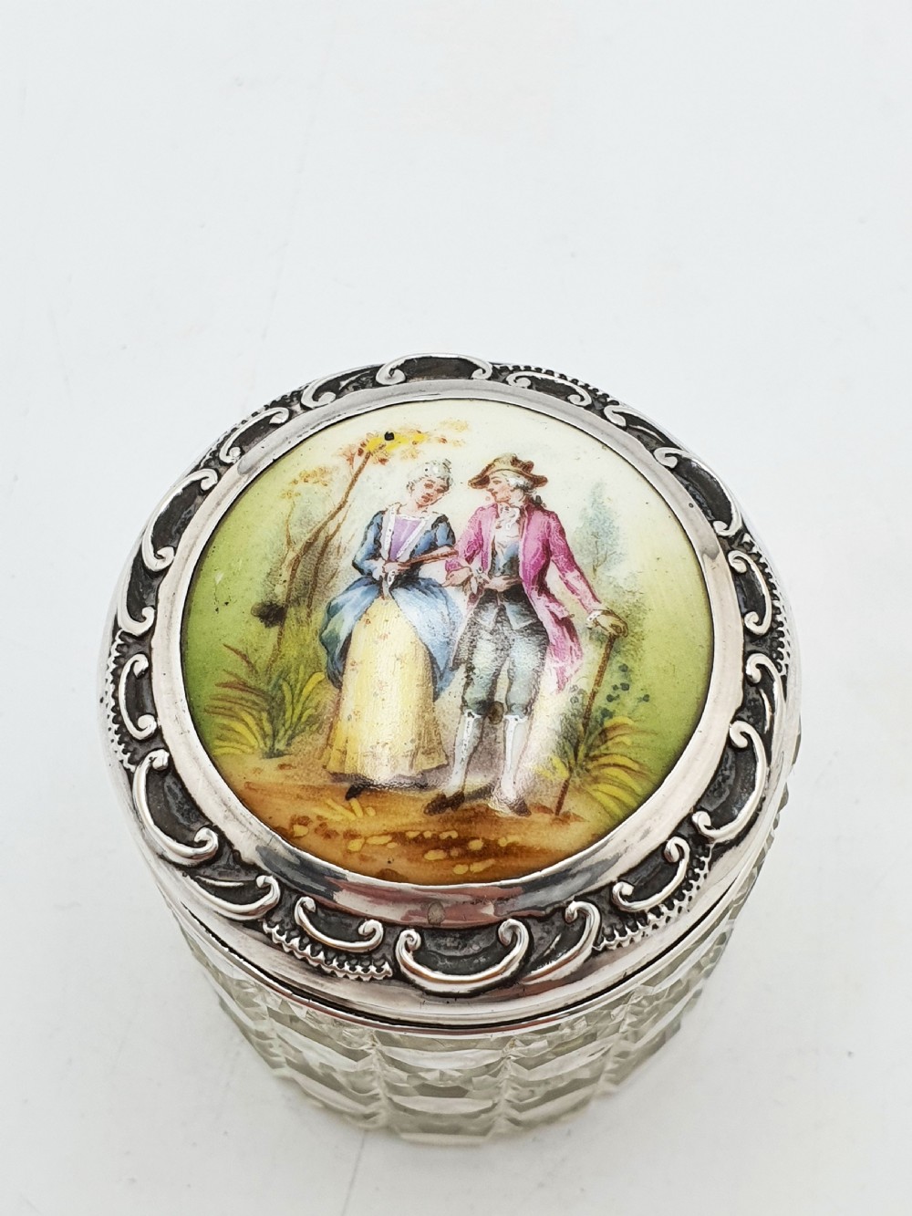 An Edwardian round cut-glass vanity jar featuring a sterling silver mounted lid inset with a hand-painted porcelain plaque depicting an eighteenth-century courting couple.