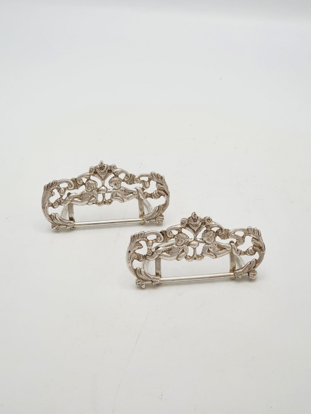 A pair of Victorian sterling silver place card holders featuring detailed cherubs framed by pierced scrolling foliage.