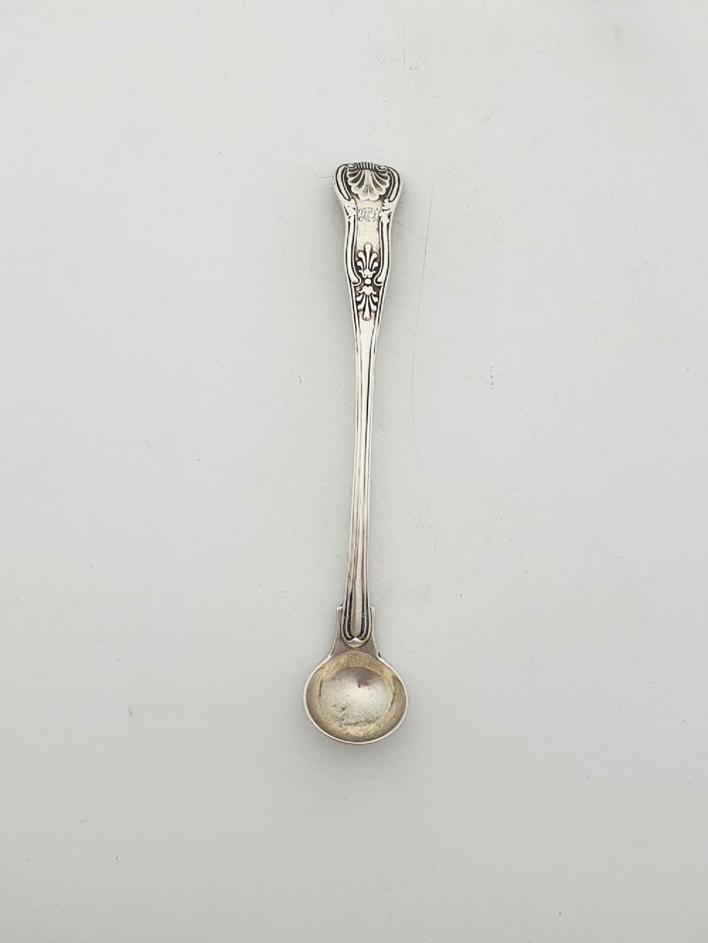 An antique sterling silver Kings pattern condiment spoon featuring a relief shell motif handle and a small rounded bowl.