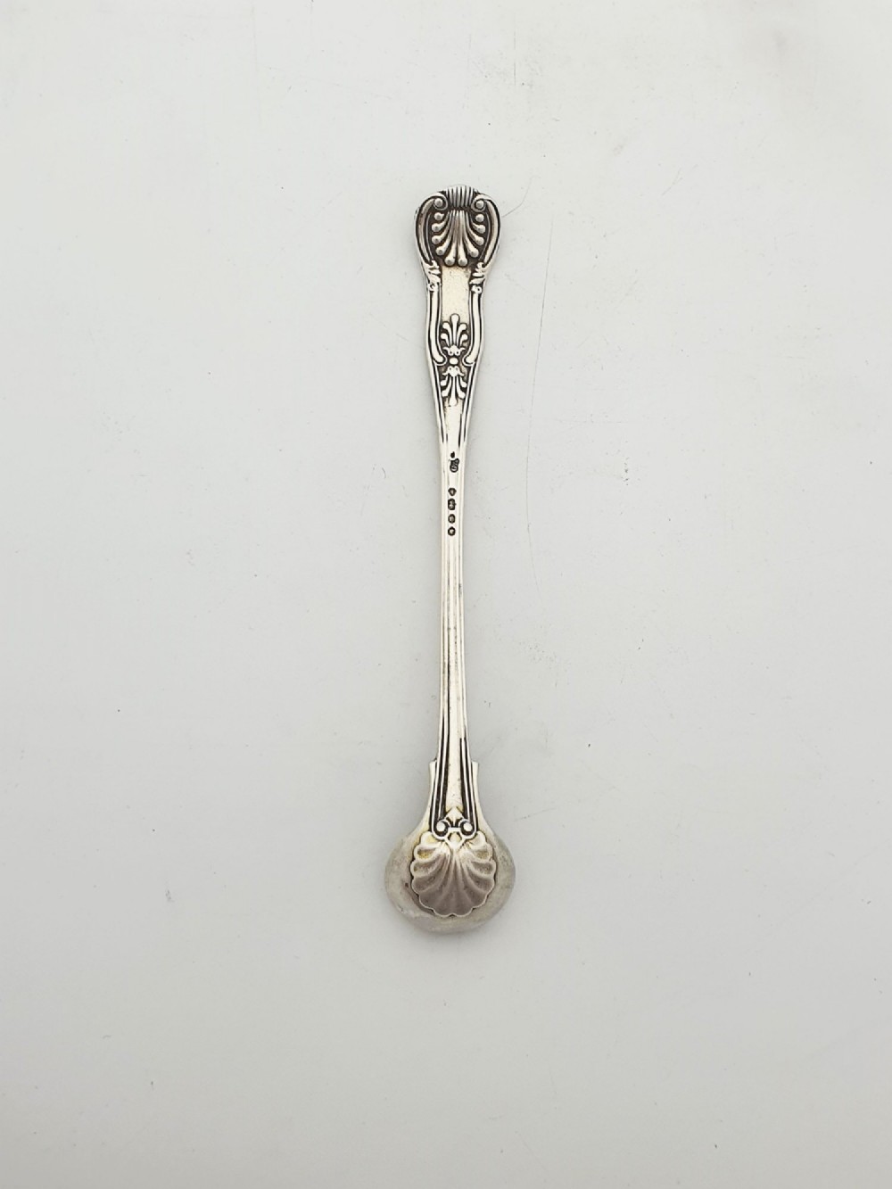 An antique sterling silver Kings pattern condiment spoon featuring a relief shell motif handle and a small rounded bowl. View II
