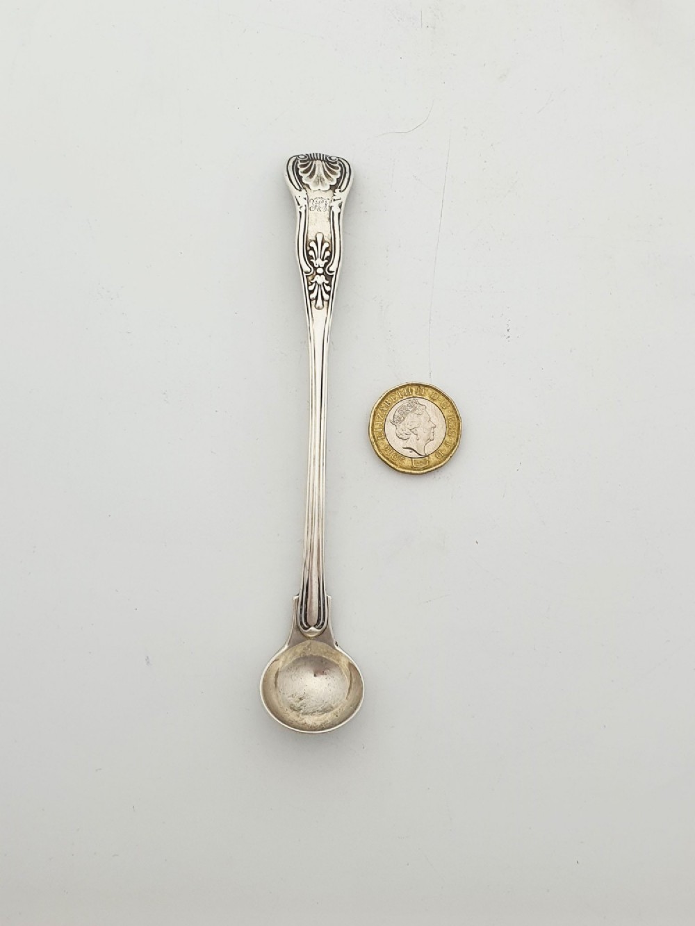 An antique sterling silver Kings pattern condiment spoon featuring a relief shell motif handle and a small rounded bowl. View IV