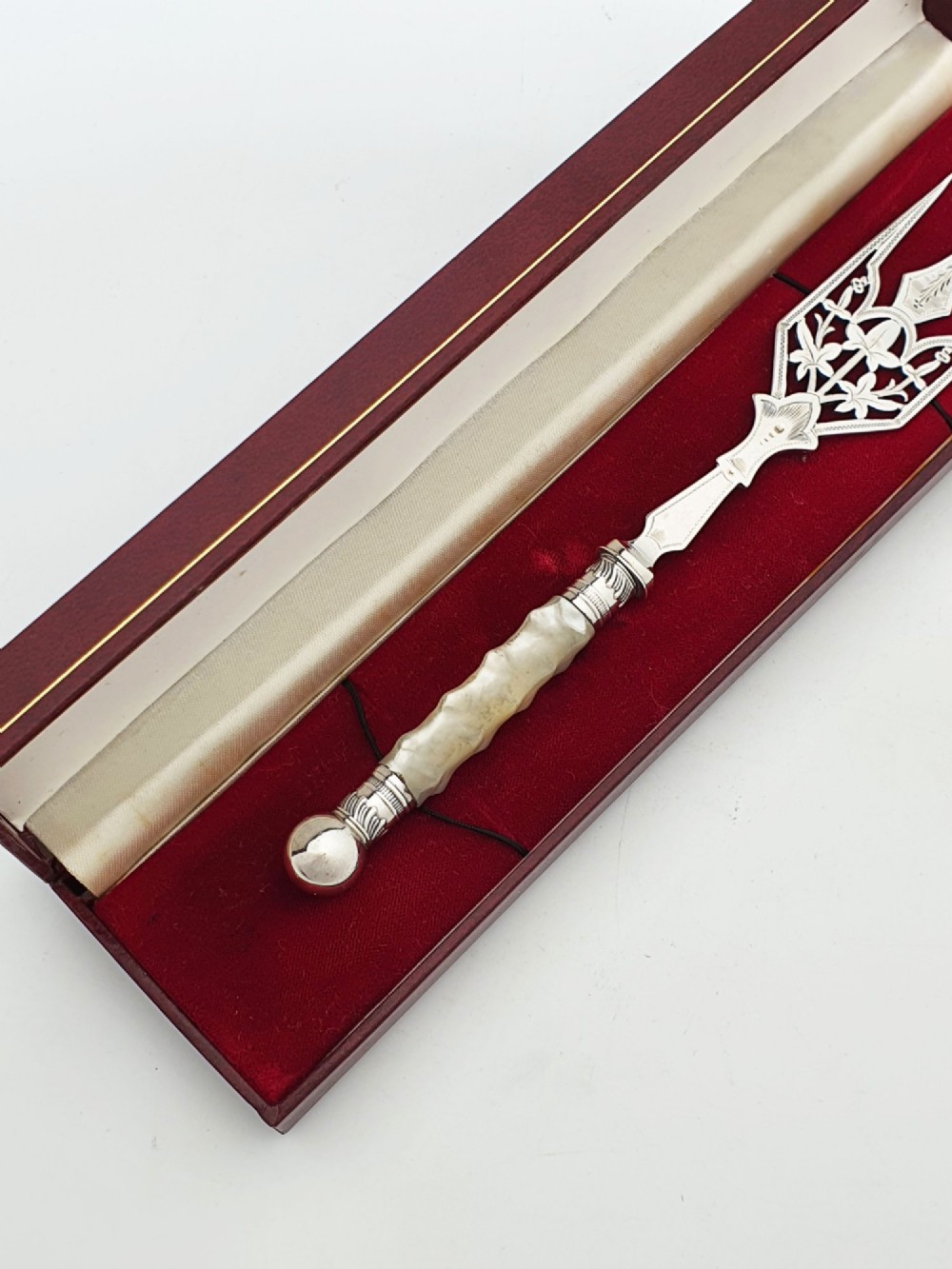 An Edwardian sterling silver bread fork featuring pierced foliage tines and a carved mother-of-pearl handle resting inside a red fitted box. View III