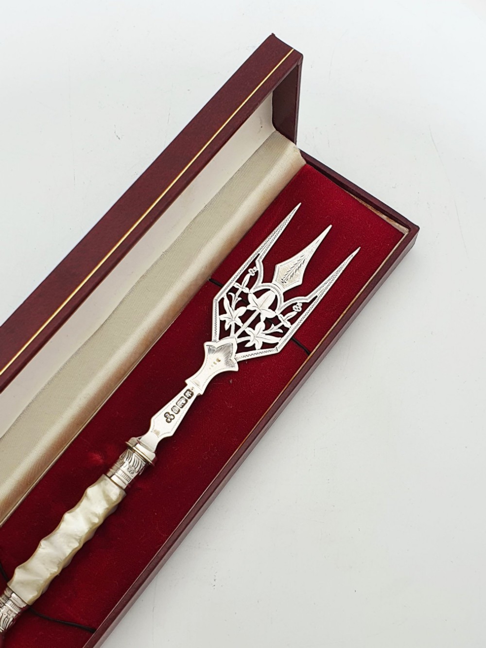 An Edwardian sterling silver bread fork featuring pierced foliage tines and a carved mother-of-pearl handle resting inside a red fitted box. View IV