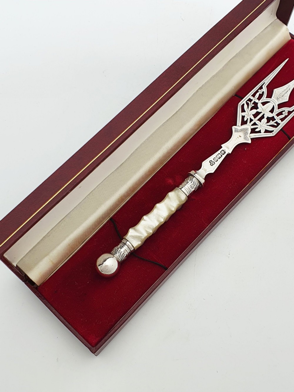 An Edwardian sterling silver bread fork featuring pierced foliage tines and a carved mother-of-pearl handle resting inside a red fitted box. View V