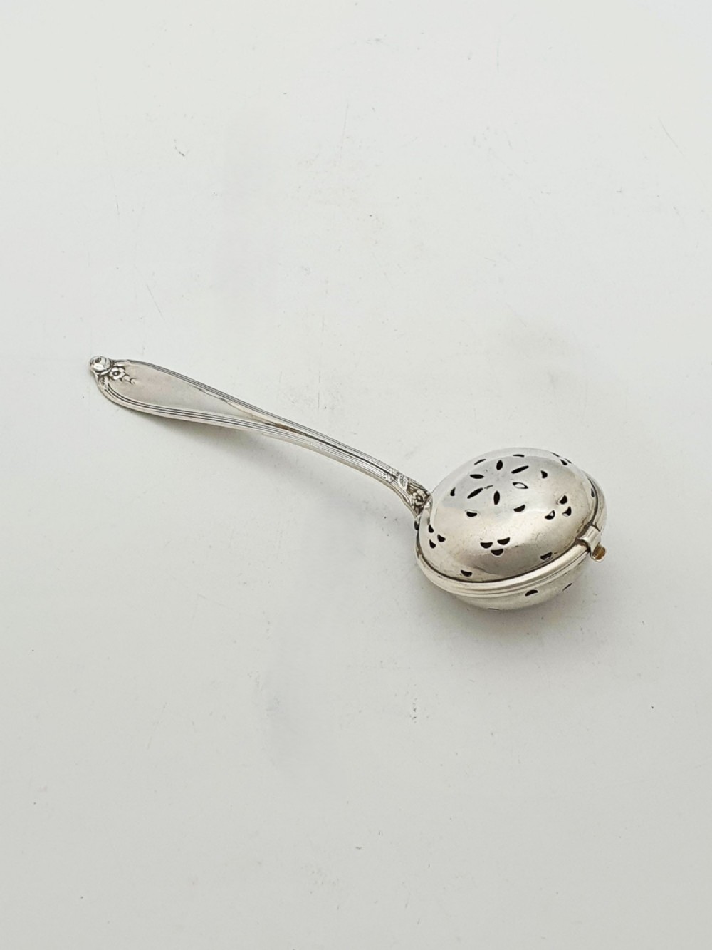 A George V sterling silver hinged tea infuser spoon featuring a perforated spherical bowl attached to a decorated handle.